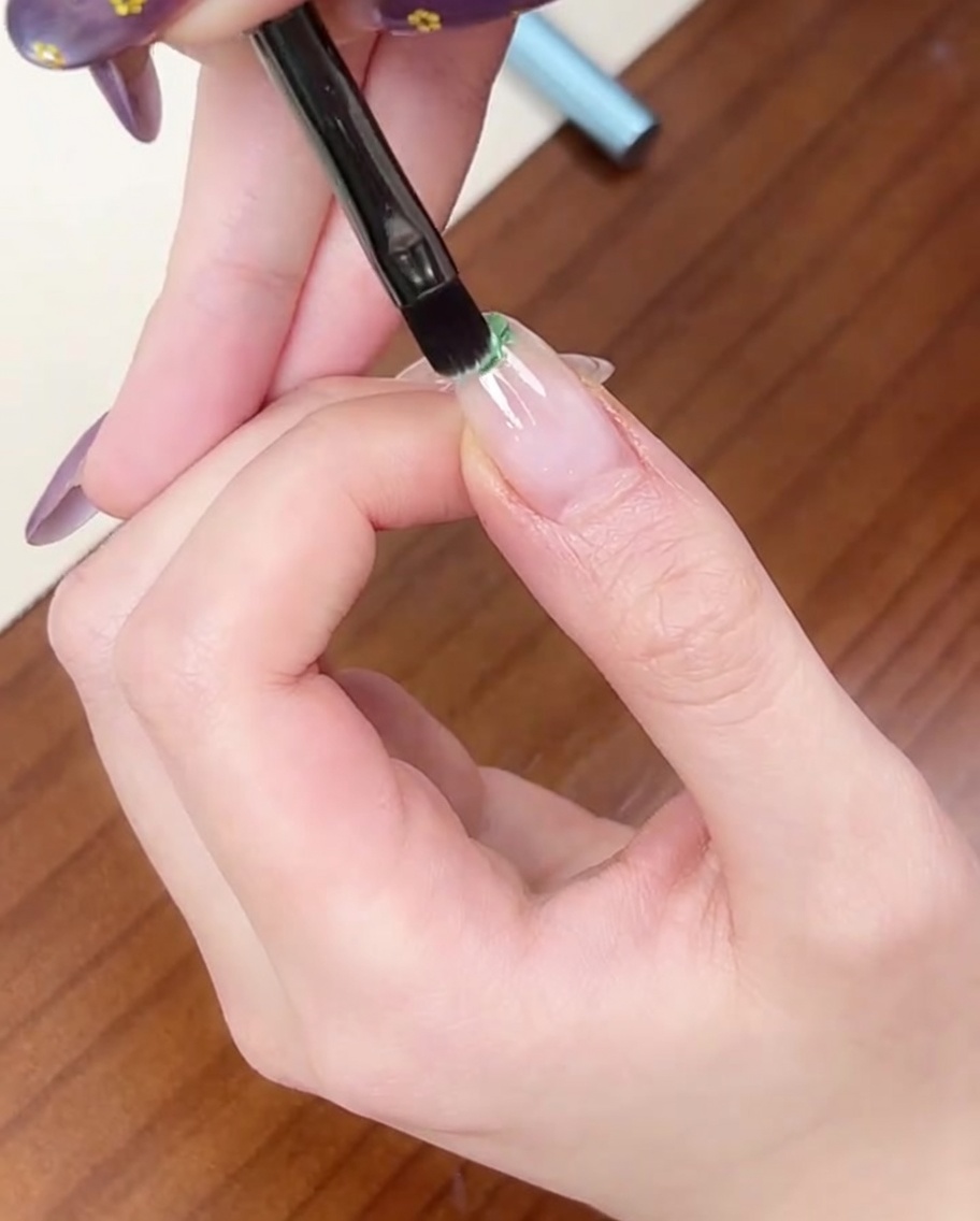 Un pincel plano aplicando una pequeña gota de esmalte en gel verde claro en la punta de una uña natural.