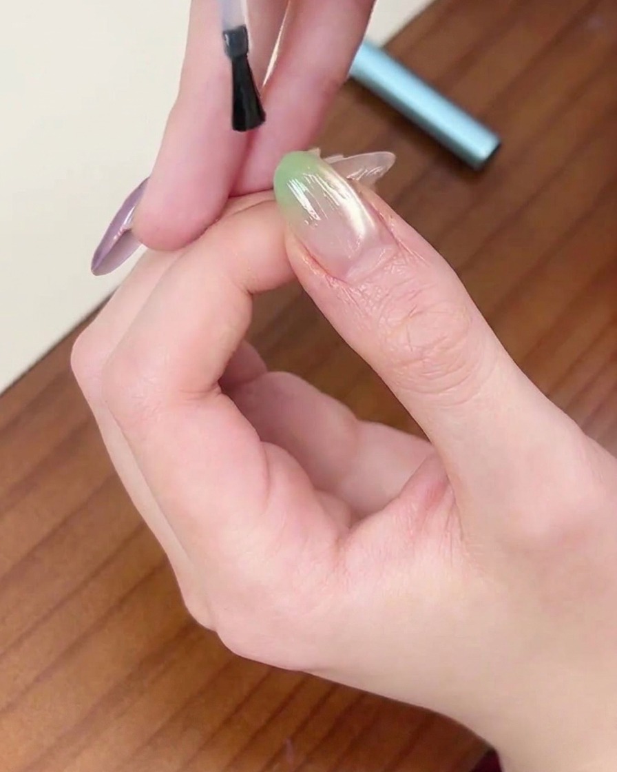 Un pincel aplicando una capa final de top coat transparente sobre una uña con degradado verde.