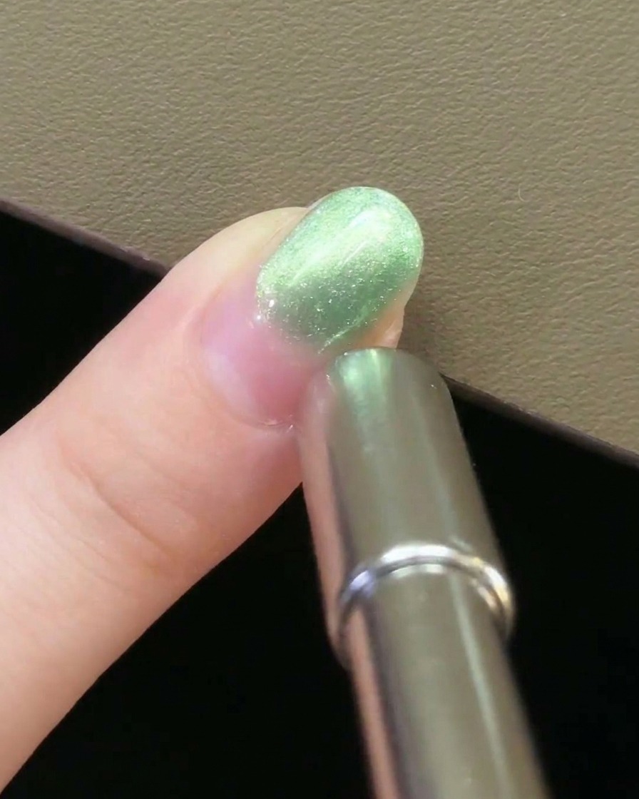 Un imán metálico para uñas sostenido paralelamente al lado de una uña cubierta con gel magnético verde.