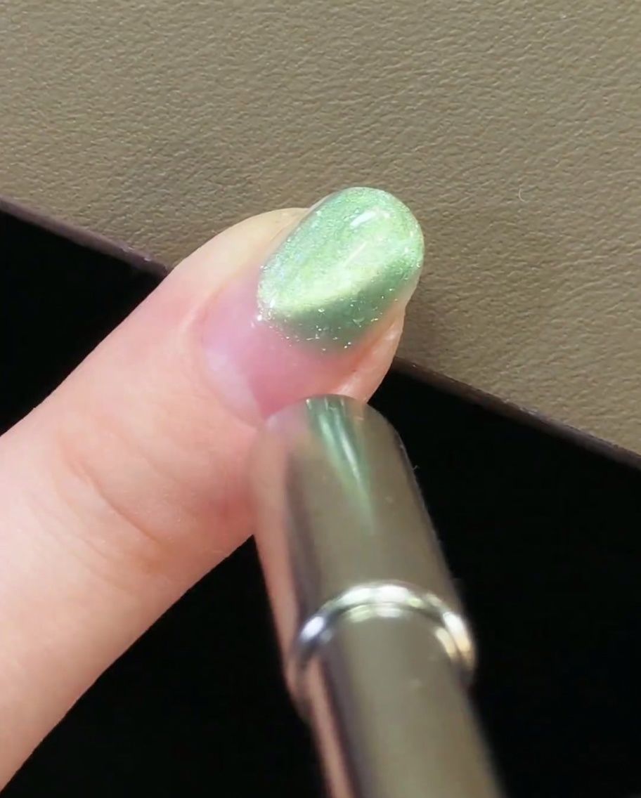 Un imán metálico para uñas posicionado en la zona de la cutícula de una uña con gel magnético ojo de gato verde.