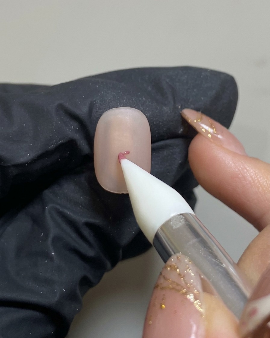 Una herramienta de silicona colocando un pequeño punto de gel rosa en el centro de una uña con base mate.