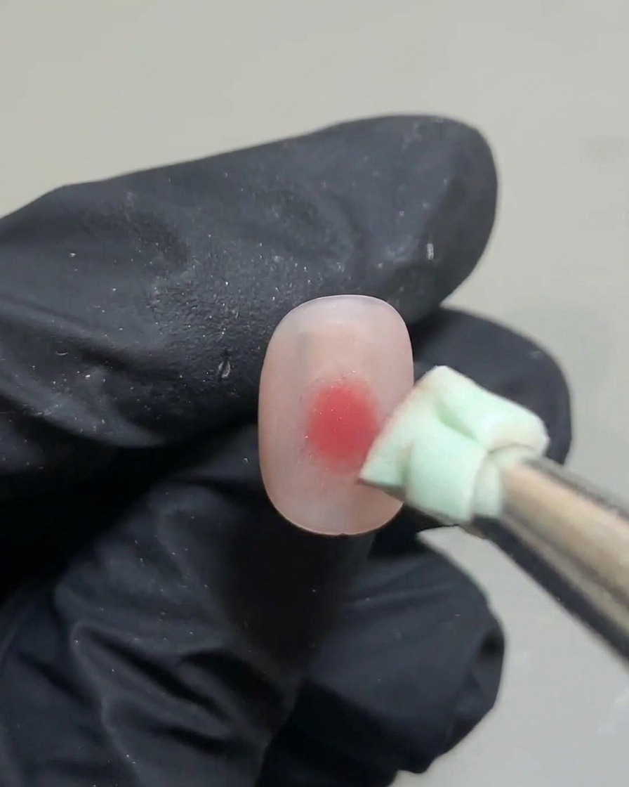 Unas pinzas sosteniendo un pequeño trozo de esponja verde, dando toques sobre un punto de gel rosa en una uña.