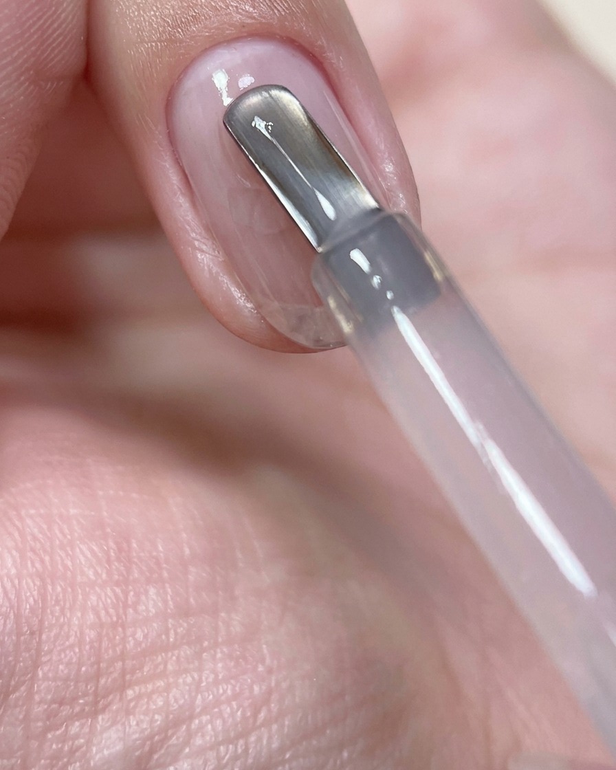 Primer plano de un pincel de uñas aplicando una perla de gel de construcción rosa traslúcido en el centro de una uña natural preparada.