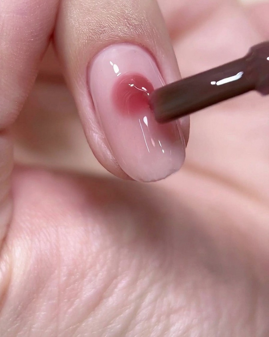 Un pincel de uñas depositando una perla precisa y redonda de gel color rosa vino profundo en el centro de una base de uña rosa claro.