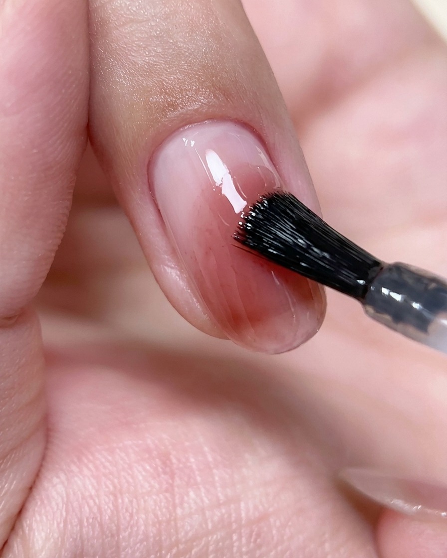 Un pincel de uñas aplicando una capa fina de top coat transparente sobre un diseño de uña blush rosa.