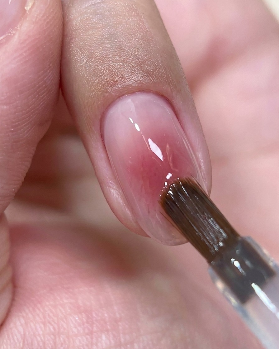 Un pincel de uñas depositando una pequeña perla de gel top coat transparente sobre el centro de una uña rosa brillante.