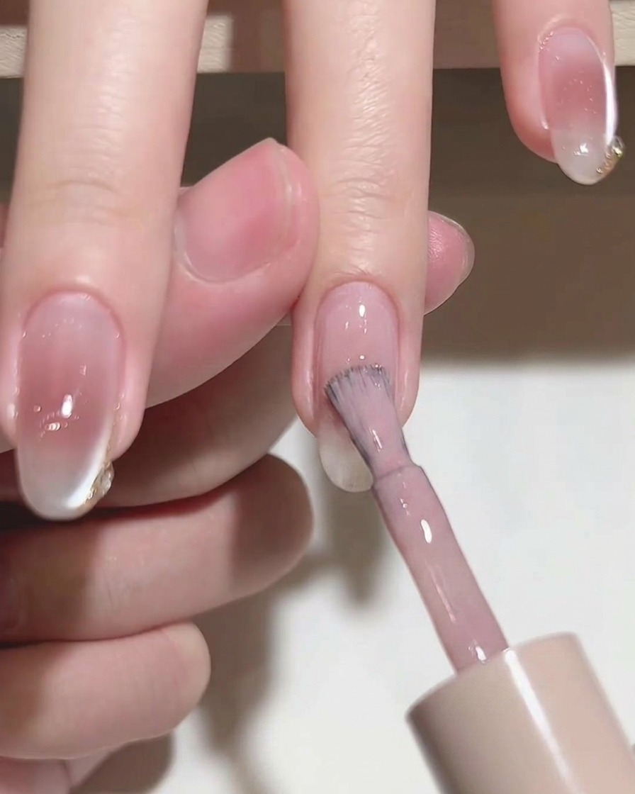 Primer plano de un pincel aplicando esmalte en gel rosa translúcido sobre una uña natural.