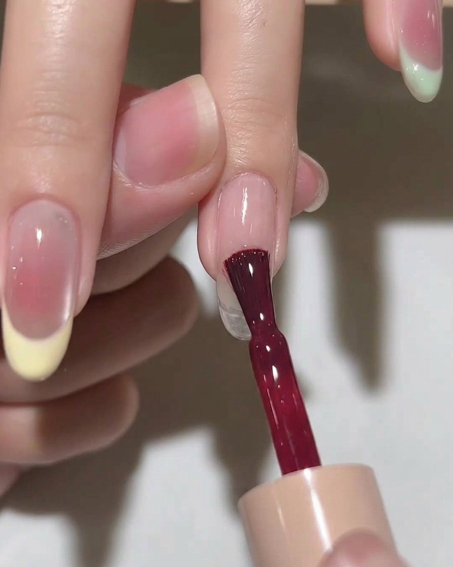 Un pincel aplicando una capa de esmalte en gel rosa translúcido sobre una uña almendrada.