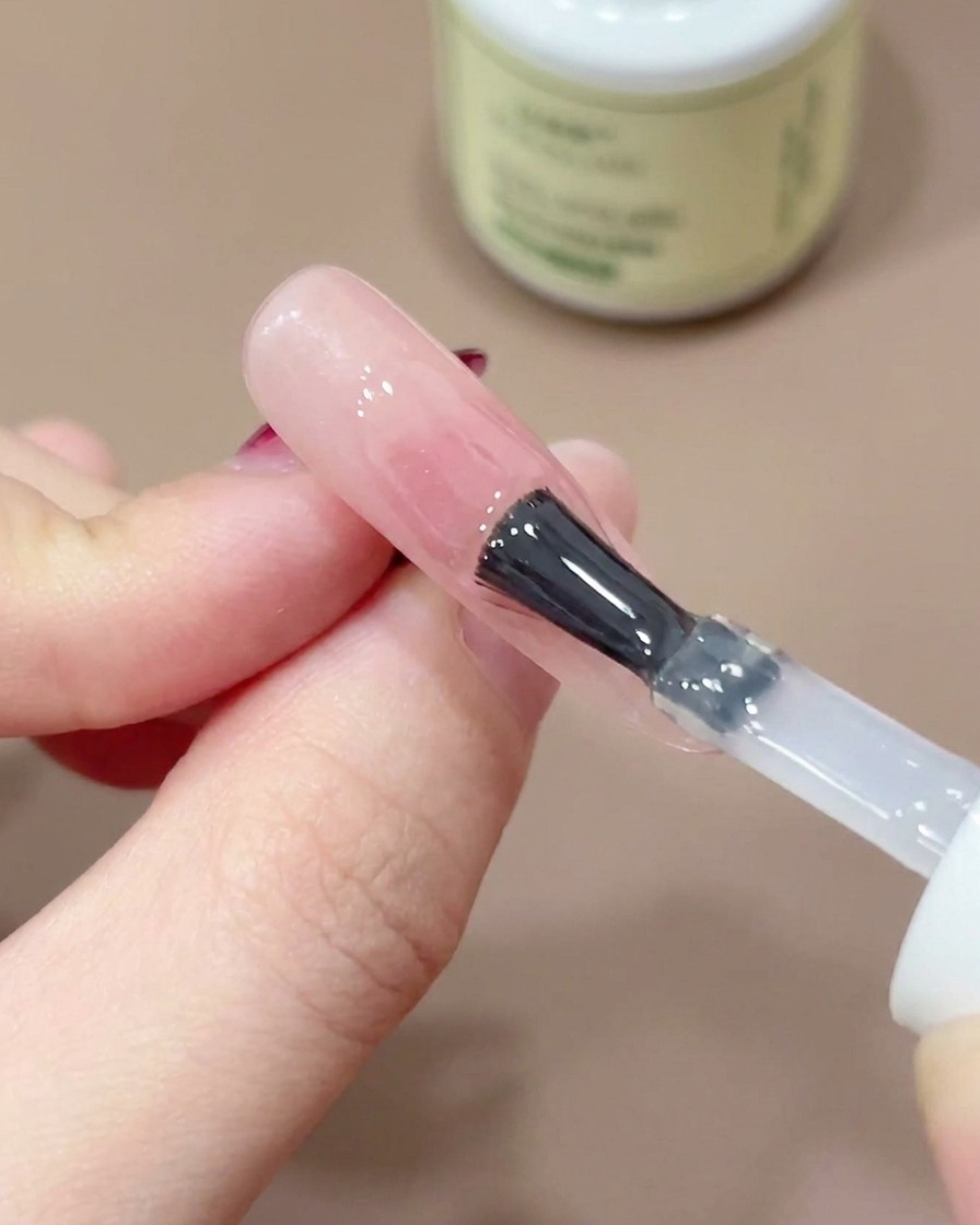 Un pincel de uñas aplicando un top coat transparente y brillante sobre un diseño blush rosa.