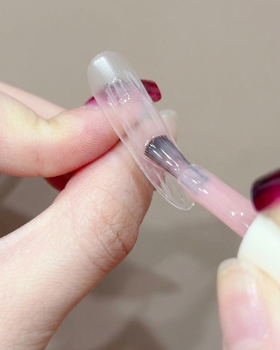 Primer plano de un pincel de gel aplicando una pequeña gota de gel durazno en el centro de una uña sin esmalte.