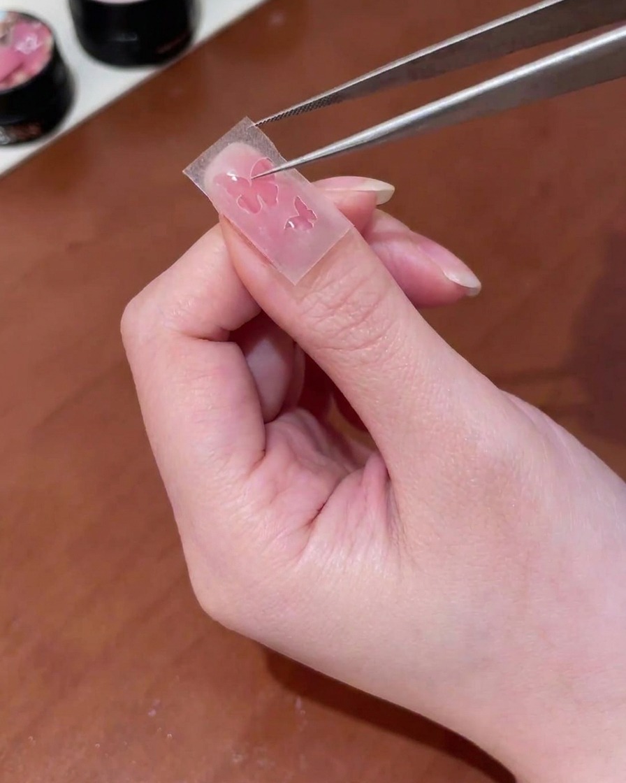 Pinzas colocando una pegatina con forma de mariposa sobre la superficie de la uña.
