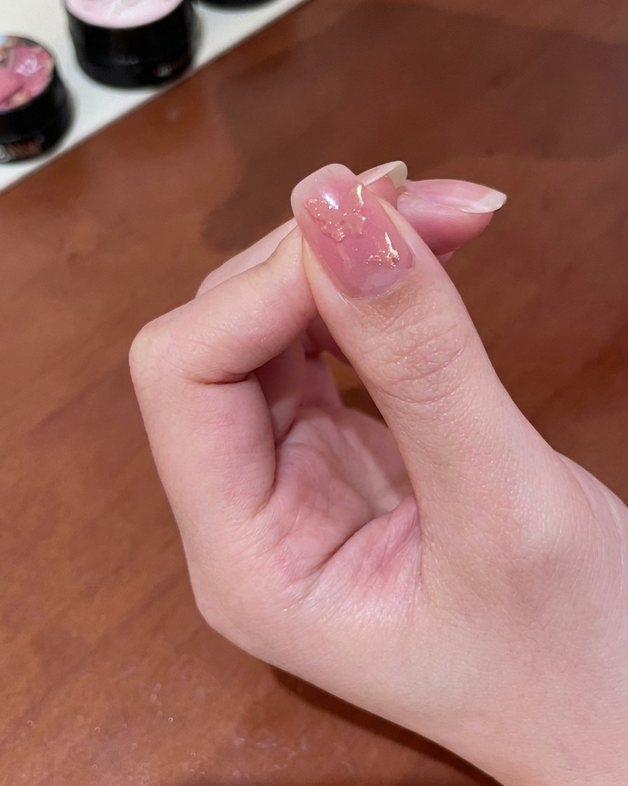 Primer plano de una mano sosteniendo un pequeño imán cilíndrico sobre una uña recubierta con gel magnético dorado húmedo, revelando un brillo cambiante.