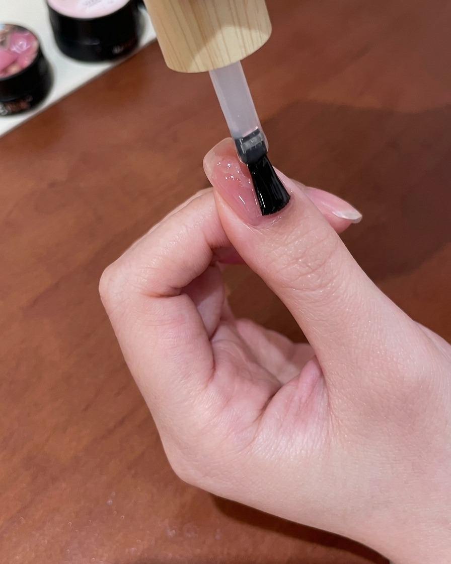 Un pincel aplicando una capa de top coat de gel transparente y brillante sobre una uña rosa nude terminada que presenta un diseño de mariposa ojo de gato dorado.