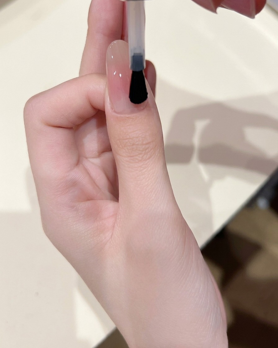 Un pincel de uñas aplicando una capa fina de gel base transparente y brillante sobre una uña que tiene un centro blush suave.