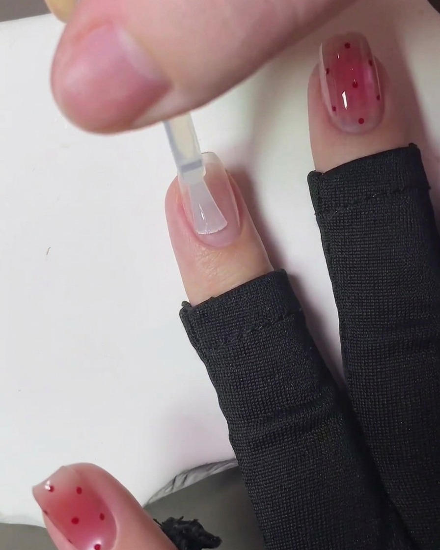 Un pincel aplicando un top coat transparente y brillante sobre el diseño terminado de uñas strawberry blush con lunares.