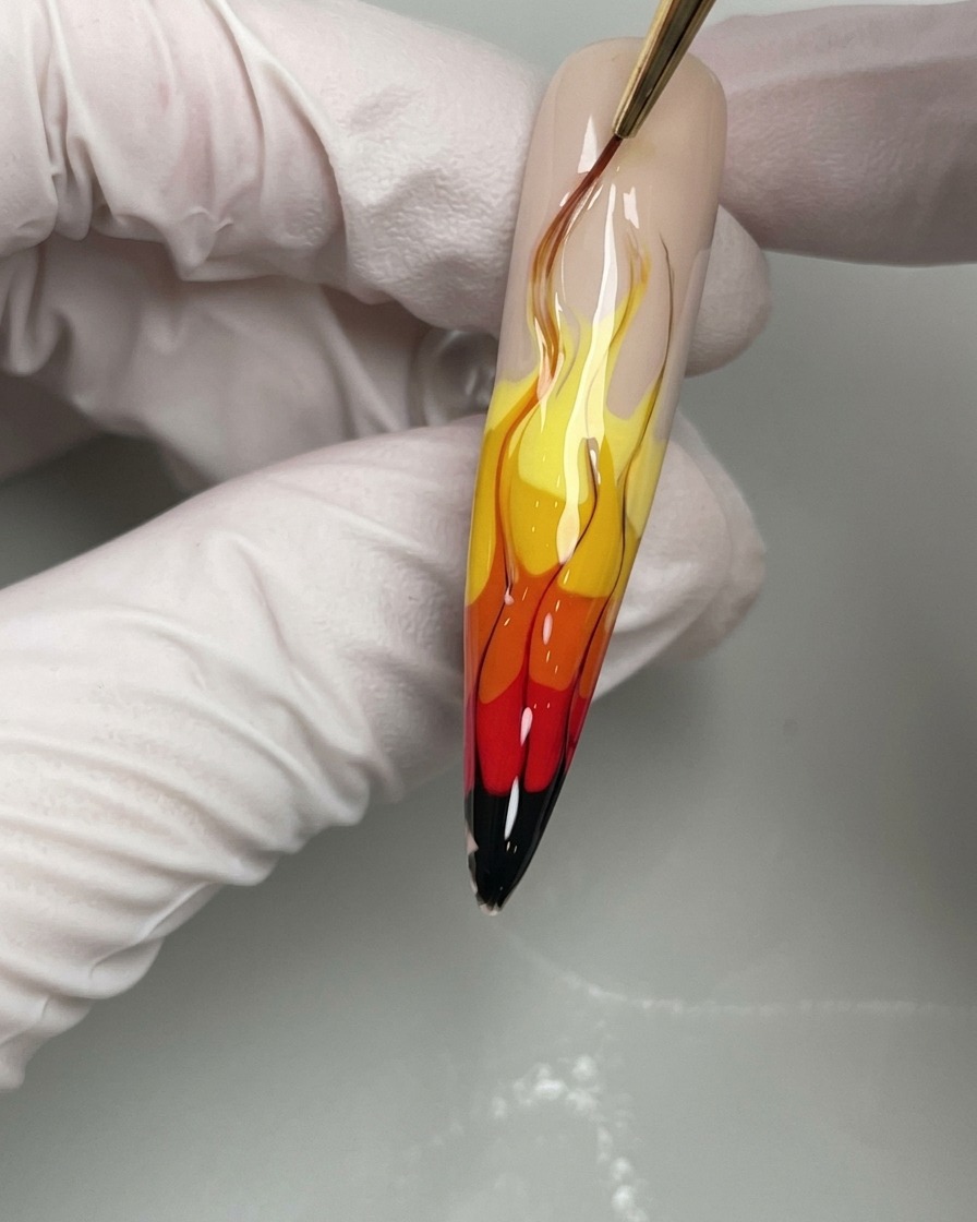 Primer plano de un pincel de uñas aplicando líneas finas de gel negro para definir los bordes de un diseño de uñas con llamas amarillas, naranjas y rojas.