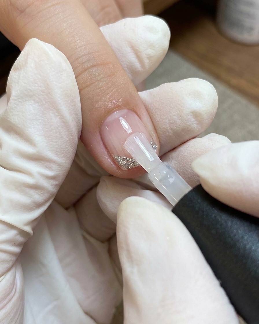 Un pincel aplicando una capa de top coat transparente sobre una uña decorada con un corazón y líneas finas blancas y plateadas.