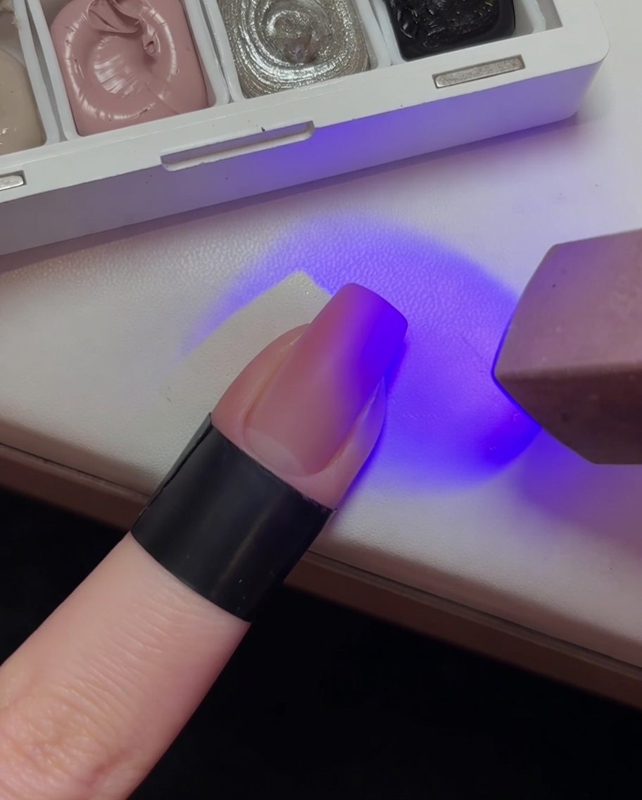 Una uña con base degradada rosa claro siendo curada bajo la luz de una lámpara UV morada.