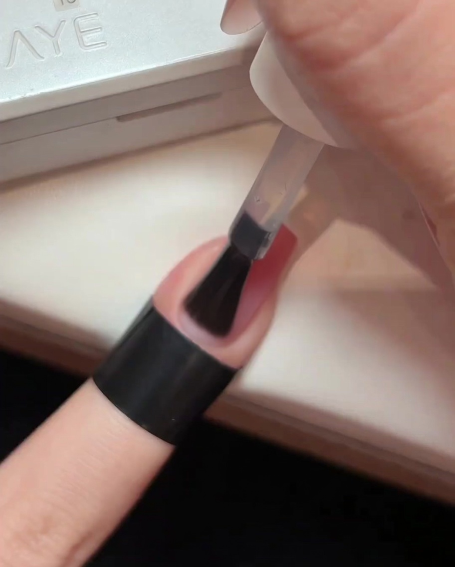 Un pincel aplicando una capa de top coat transparente brillante sobre una uña degradada rosa.