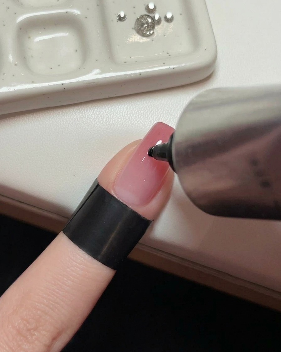 Una mano sosteniendo un tubo metálico exprimiendo una gota de gel adhesivo sobre el centro de una uña rosa brillante.
