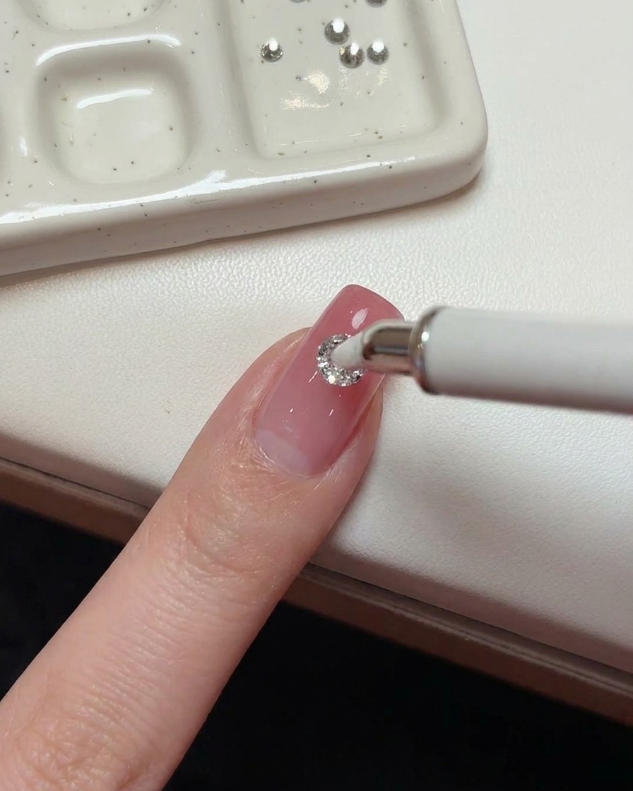 Un lápiz con punta de cera blanca colocando un cristal redondo grande de 5mm en el centro de una uña rosa.