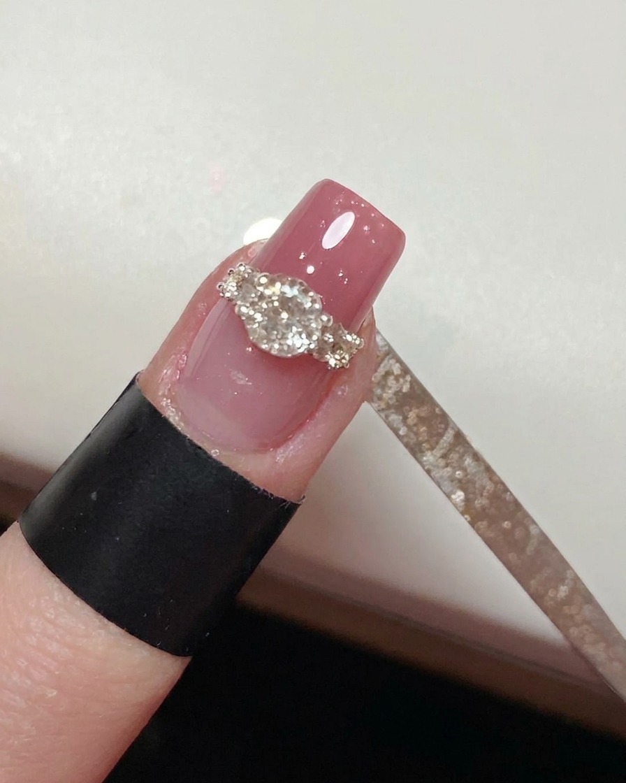 Un pincel liner fino aplicando cuidadosamente top coat alrededor de la base de un diseño de anillo de cristal completado en una uña rosa.