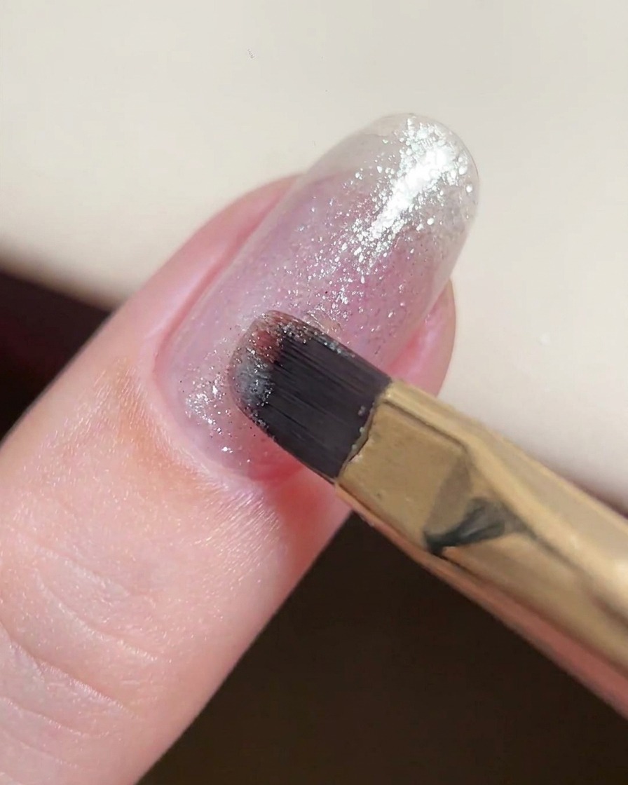 Un pincel de gel aplicando una capa fina y consistente de gel con purpurina plateada sobre una uña natural.