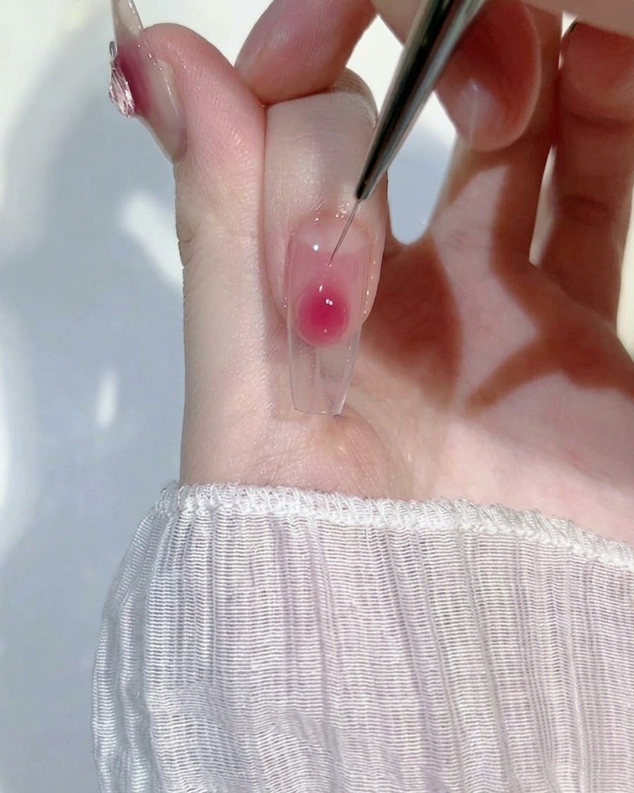 Primer plano de un pincel liner fino difuminando suavemente una gota de gel rubor rosa sobre una uña transparente para crear un efecto degradado.