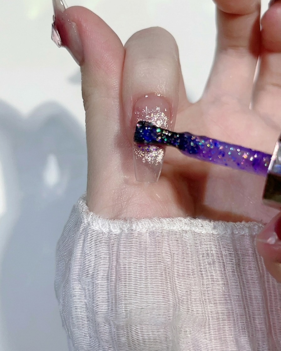 Una capa fina de gel con glitter con tintes morados siendo aplicada sobre un diseño de uñas rosa ojo de gato existente.