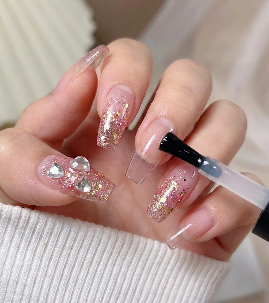 Un pincel aplicando una capa final de top coat brillante sobre una uña decorada con glitter rosa y cristales de corazón.