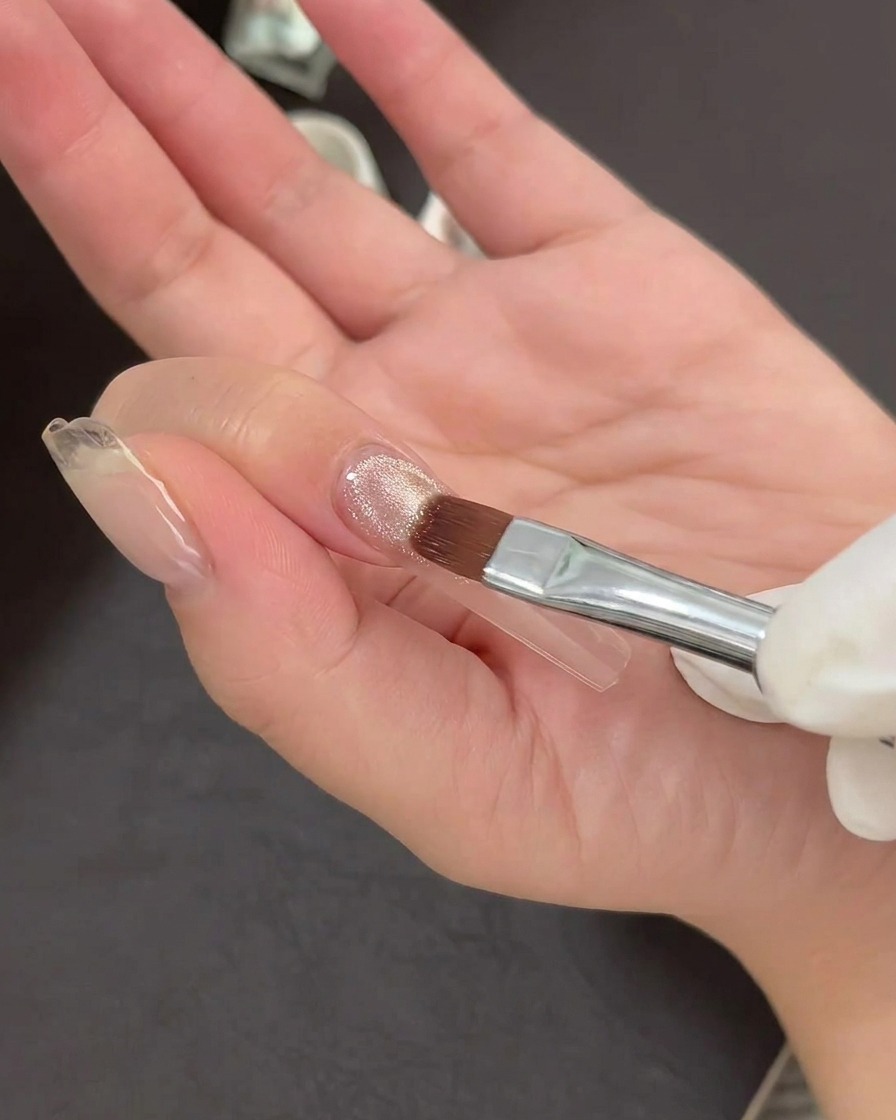 Un pincel de arte de uñas aplicando una capa fina de esmalte de gel ojo de gato brillante sobre una punta de uña transparente.