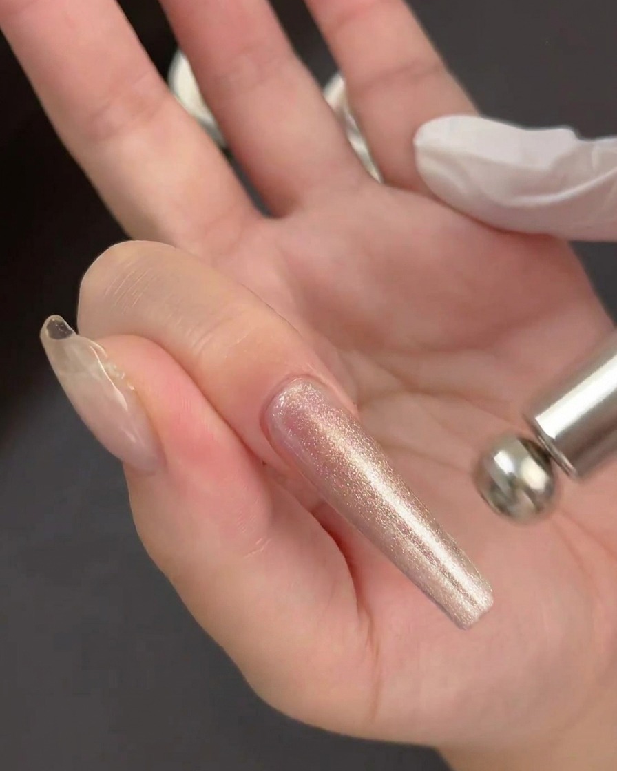 Una mano sosteniendo una herramienta magnética cilíndrica flotando justo por encima de una aplicación de gel brillante y húmeda en una extensión de uña transparente.