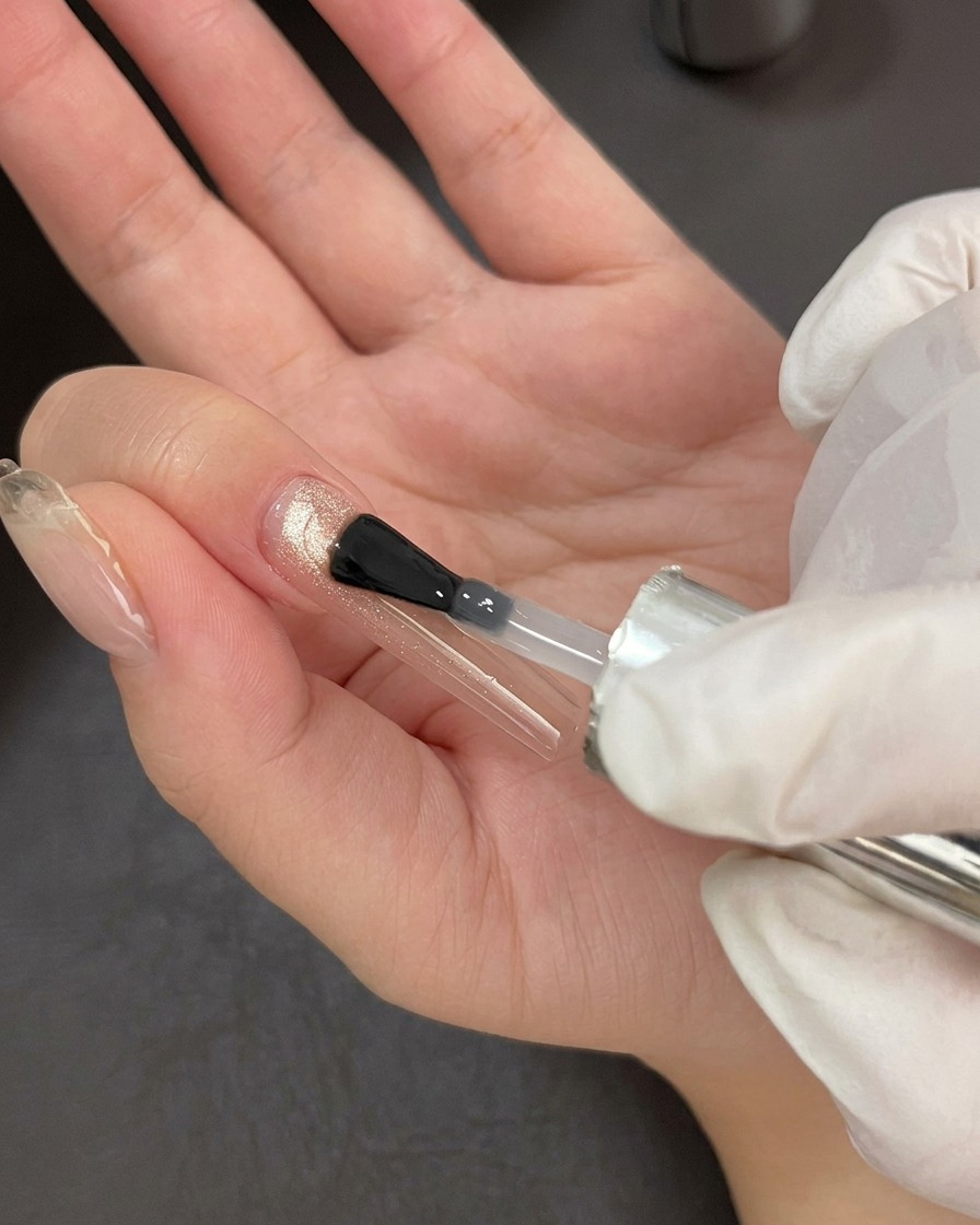 Un pincel de uñas profesional aplicando un top coat transparente y brillante sobre una base de gel ojo de gato.