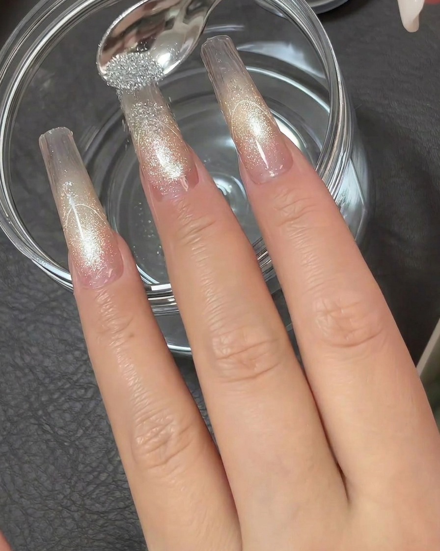 Una pequeña cuchara metálica esparciendo polvo de diamante plateado altamente reflectante sobre la punta adhesiva de una extensión de uña transparente.