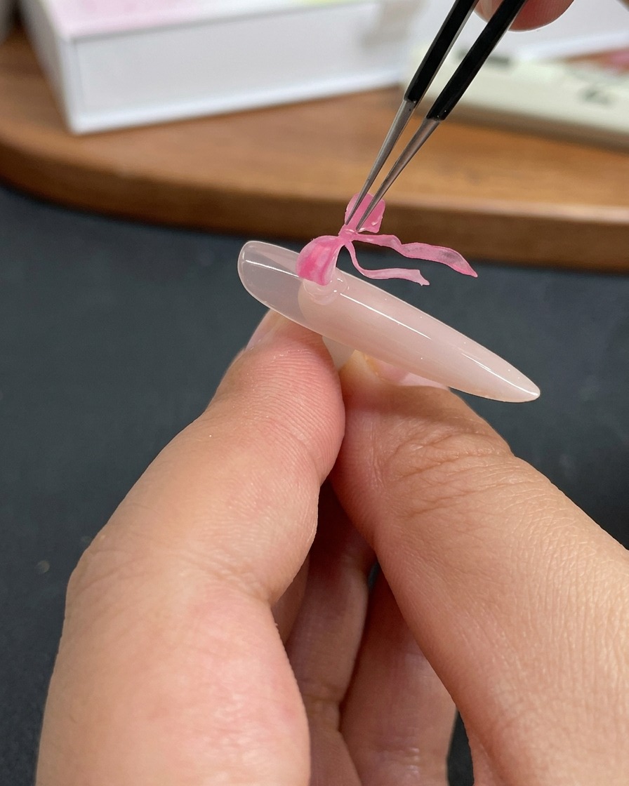 Unas pinzas colocando un delicado dije de lazo de mariposa rosa sobre una uña recubierta de gel transparente.