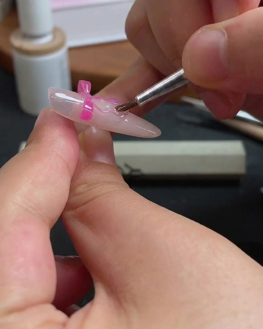 Un pincel fino aplicando con cuidado gel transparente alrededor de los bordes de un dije de lazo rosa en una uña.