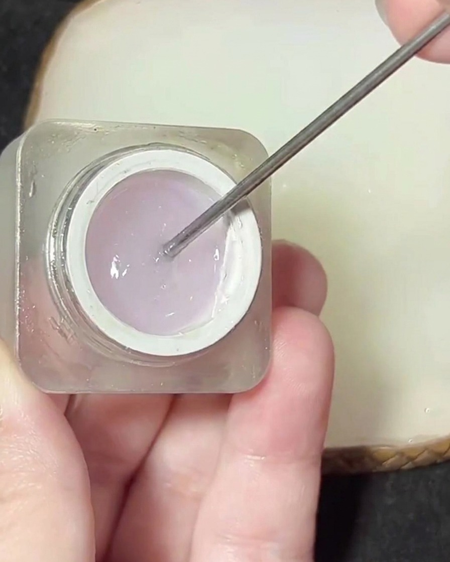 Una espátula metálica levantando un producto de gel espeso y transparente desde un pequeño frasco.