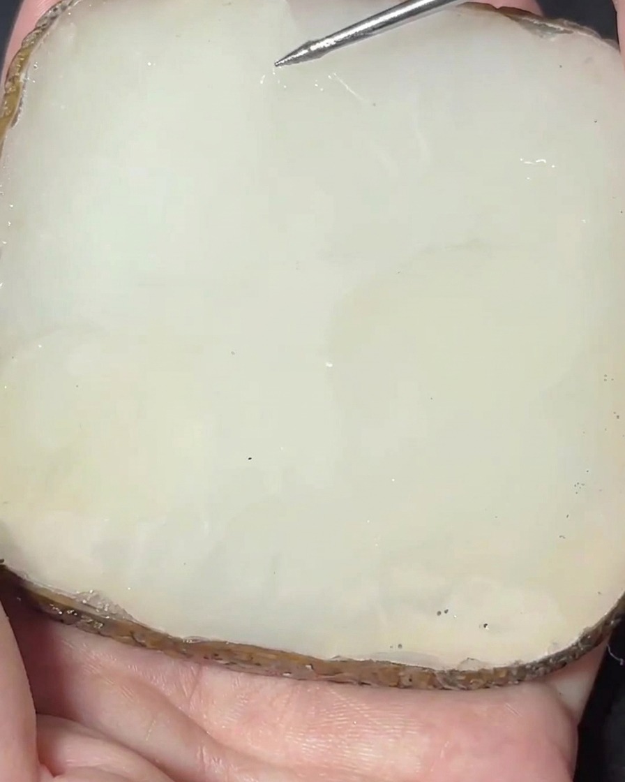 Una espátula metálica mezclando un pigmento rosa en una base de gel transparente sobre una paleta de piedra.