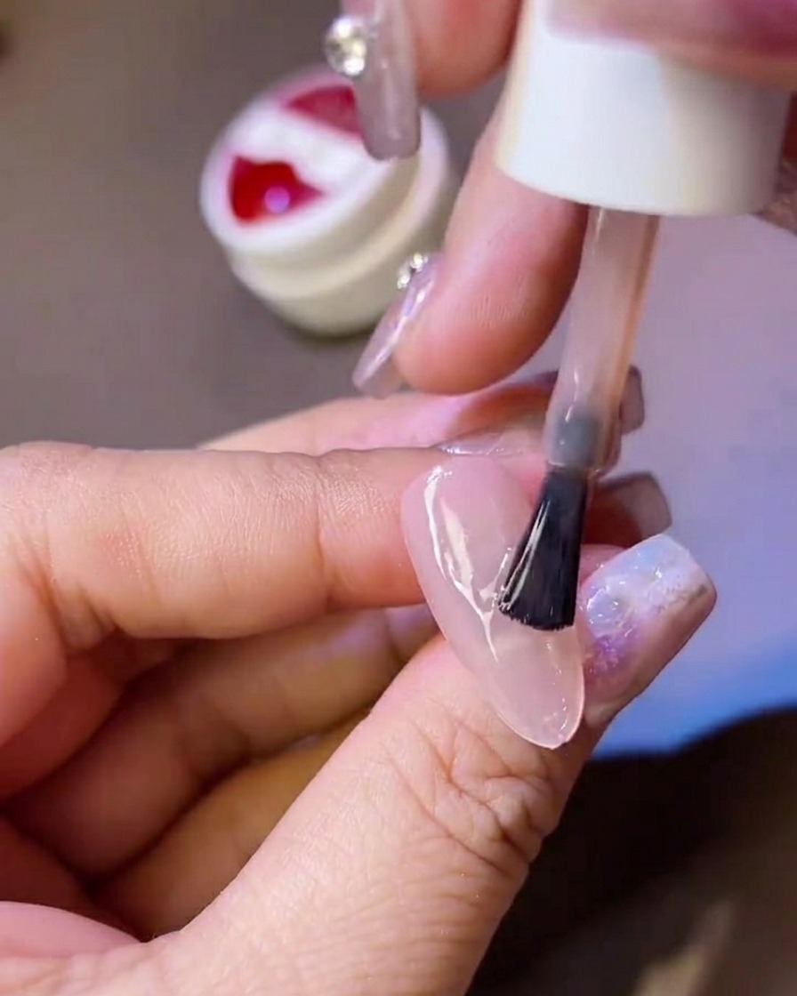 Una mano sosteniendo un pincel de esmalte, aplicando una capa de top coat mate sobre una uña postiza translúcida.