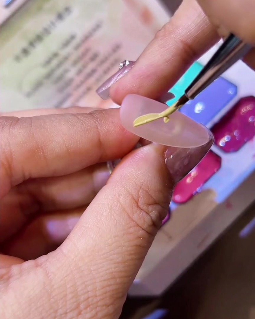 Un pincel fino delineador dibujando la forma de media ala de mariposa sobre una uña mate usando esmalte en gel amarillo.