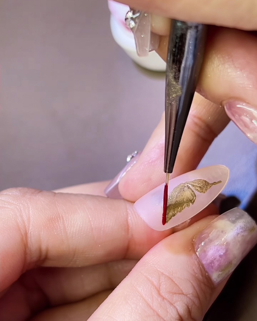 Un pincel fino aplicando un contorno de gel rojo alrededor de un ala de mariposa metálica dorada sobre una uña.