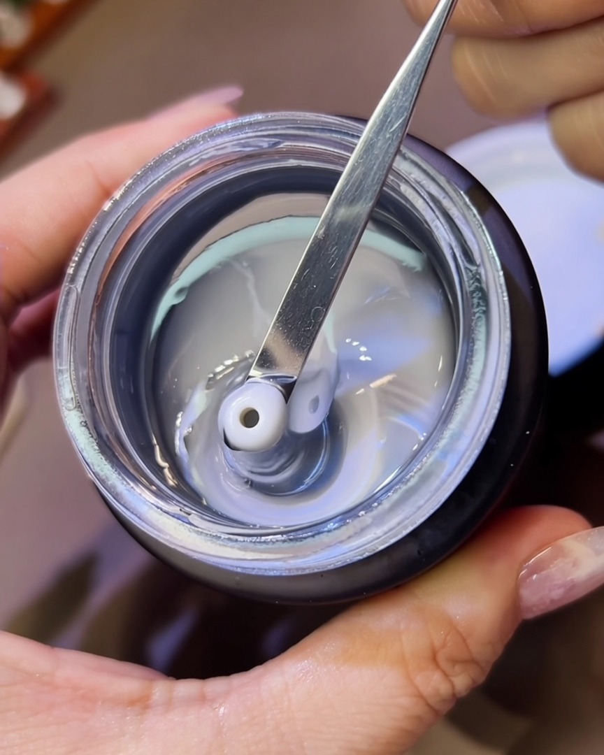 Primer plano de una herramienta de metal colocando pequeñas perlas de gel de esculpido espeso y transparente sobre una uña.