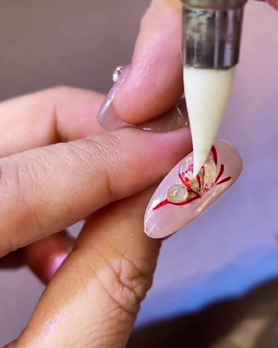 Una herramienta de silicona puntiaguda presionando y aplanando una perla de gel de esculpido transparente sobre un ala de mariposa roja y dorada en una uña.