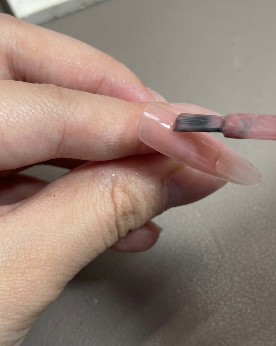 Un primer plano de un pincel aplicando gel rosa traslúcido sobre una punta de uña clara.