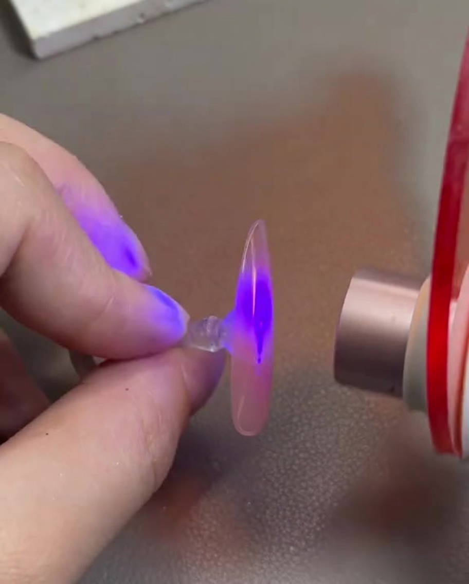 Una mano sosteniendo una uña postiza con gel rosa bajo una mini lámpara de curado UV/LED.