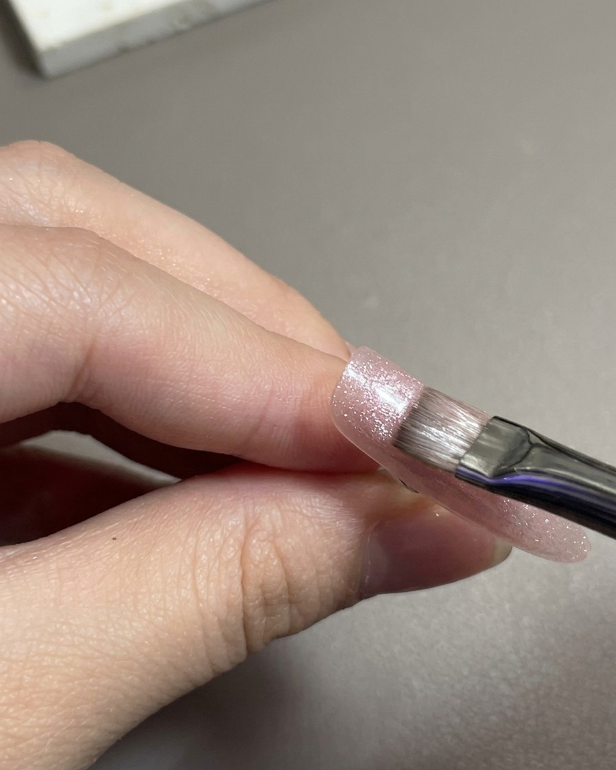 Un pincel de uñas plano extendiendo gel magnético cat eye rosa brillante sobre una uña.