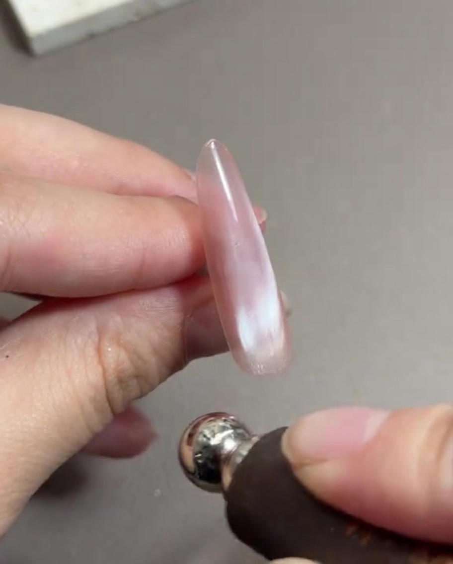 Una mano sosteniendo una herramienta magnética cilíndrica cerca de una uña larga pintada con gel rosa cat eye húmedo, moviendo las partículas magnéticas.