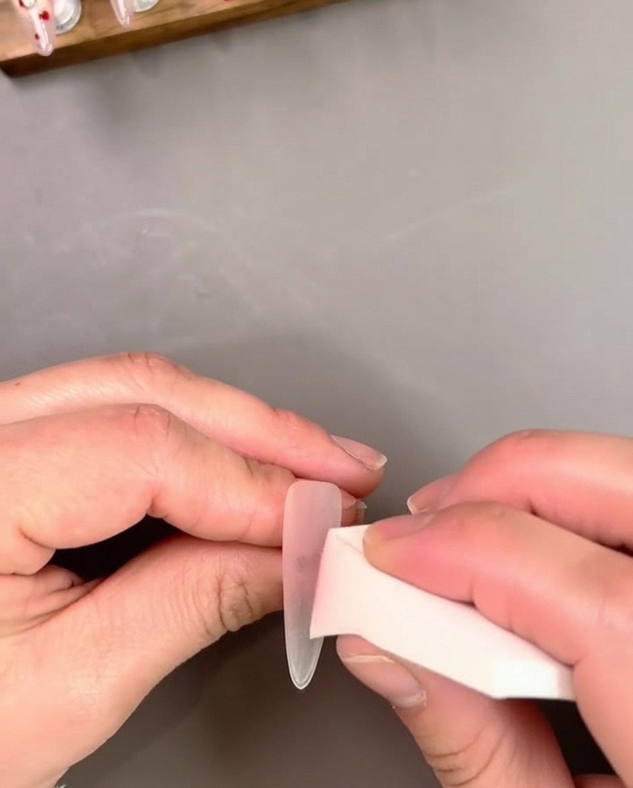 Mano sosteniendo una extensión de uña mientras aplica esmalte gel rosa traslúcido con un pincel.