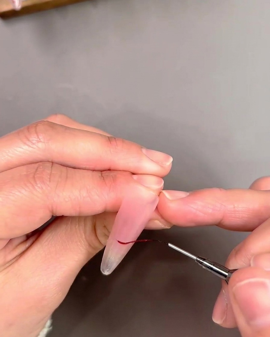 Primer plano de un pincel liner delineando una punta francesa con una línea fina de esmalte gel rojo.