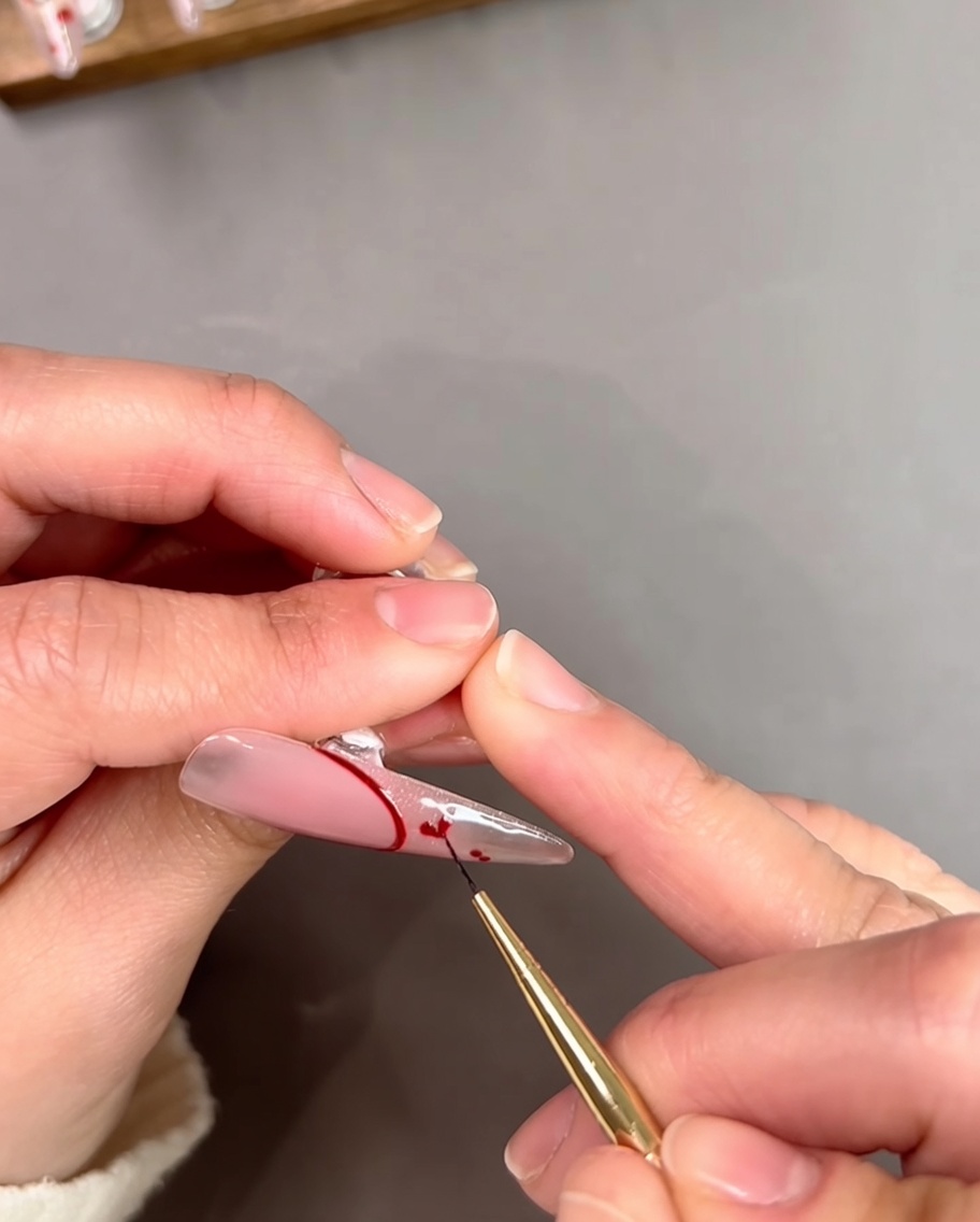 Primer plano de una manicurista usando un pincel fino para conectar dos pequeños puntos rojos en un corazón sobre una uña transparente.